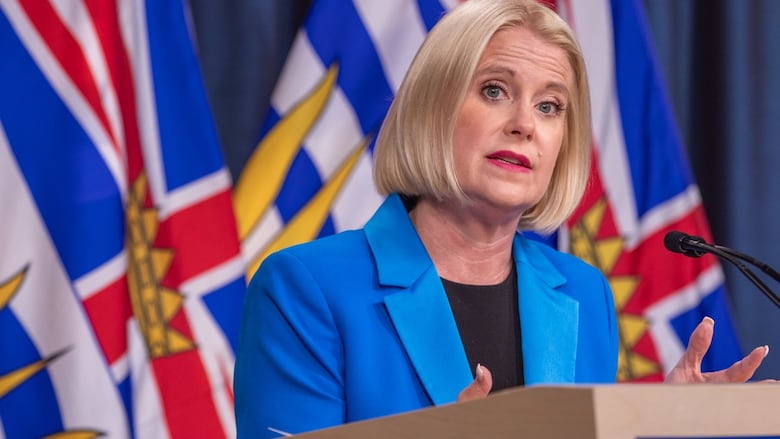 A blond woman in a blue coat speaks at a podium 