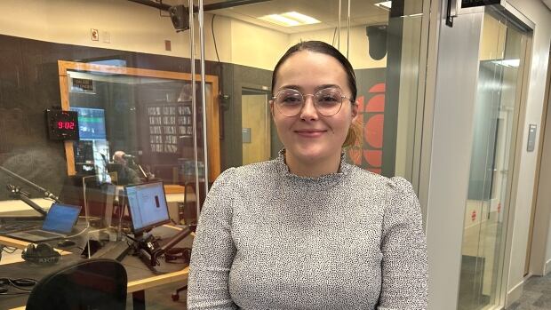 A woman wearing a grey shirt stands in front of a CBC radio recording studio.