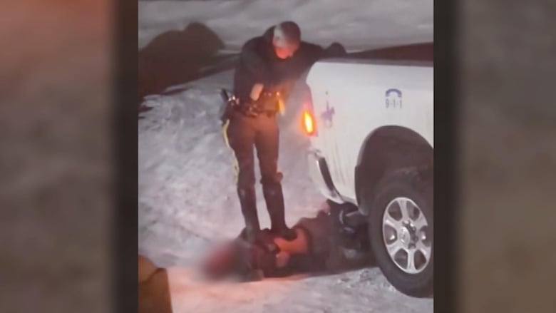A man wearing a police uniform stands on the chest of another man laying on the ground in the snow, near a white truck.