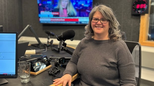 A woman with gray hair sits in a studio, next to a microphone, with a television displaying news in the background.