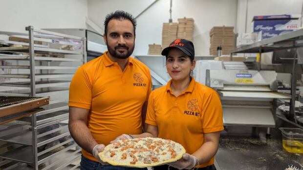 A man and woman wear matching orange polo shirts and hold a pizza.