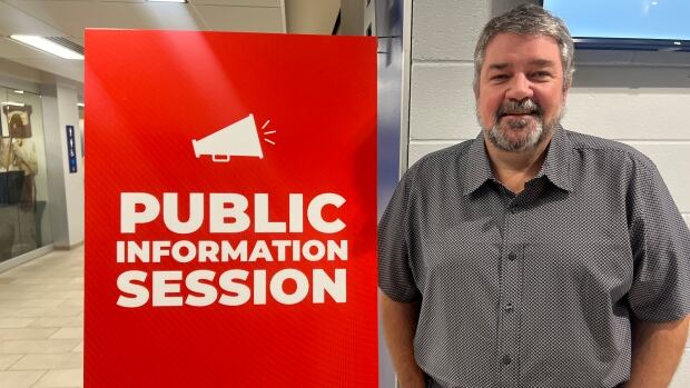 A man wearing a grey button-up shirt and jeans stands next to a red sign that says "public information session."