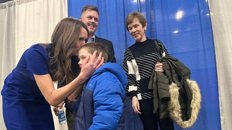 A woman in a blue dress kisses her son on the head and two other people look on. 