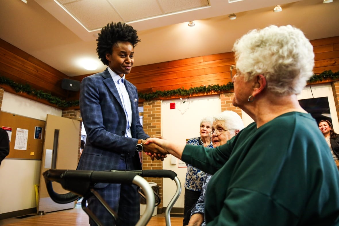A standing person in a suit shakes hands with a seated older person.