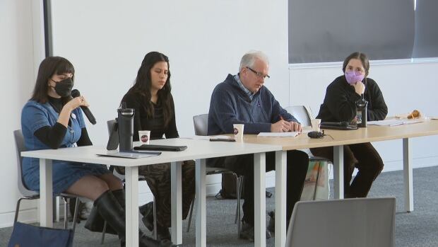 Four people sit in chairs at a table. One person holds a microphone.