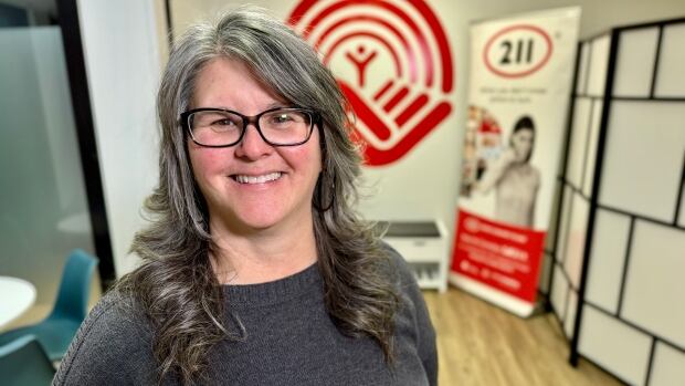 Ghislaine O'Hanley, 211 P.E.I. Educator, at the United Way office. See swag for 211 PEI. A woman wearing a dark grey sweater and glasses stands in front of a red United Way logo on a wall.