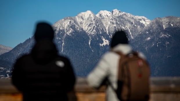 Snow on the North Shore mountains from Tatlow Park in Vancouver, B.C, on Tuesday, Feb 11, 2025. Two people are pictured in silhouette as snow-capped mountains on a clear day loom in the background.