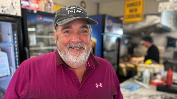 David Halfyard hasn't seen an increase in customers since the arrival of the tax break in December, but he's still convinced it had a positive impact. A man with a red Under Armour shirt and a ball cap smiles at the camera, standing in front of a counter at a diner, with refrigerators and a man at the grill behind him.
