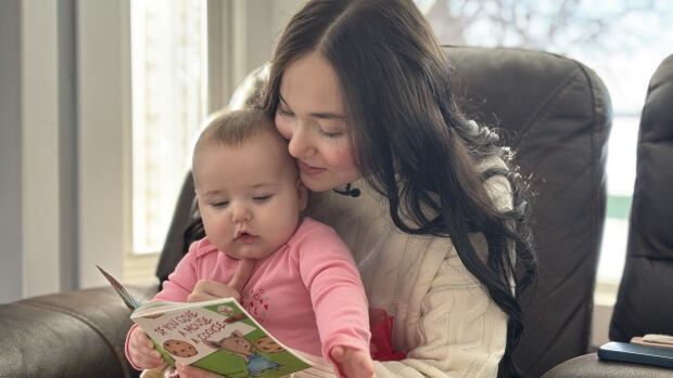 Emily Froese reads to her young daughter, Maeve A girl with long brown hair sits on a chair with a young, baby wearing a pink sweater, in her lap. She is reading her a book