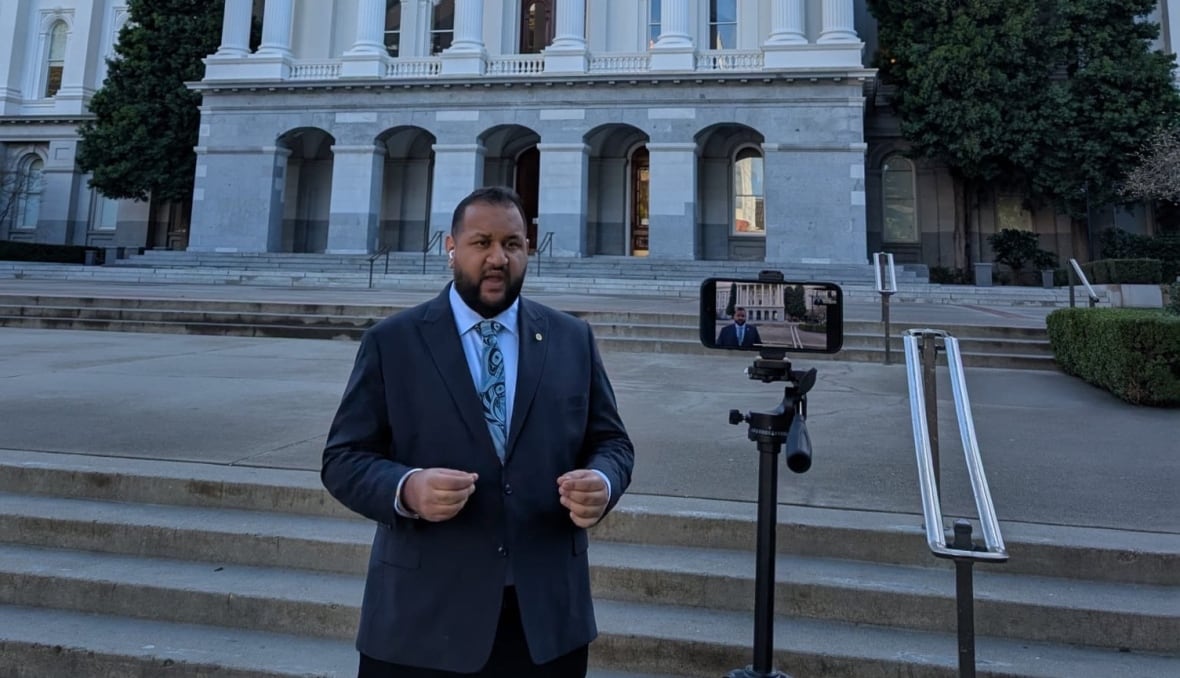 A man outside a white building is filmed by a cellphone on a tripod. 