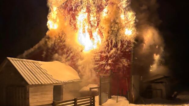 A barn on fire at night with flames lighting up the sky and a smaller snow-covered wooden structure in the foreground