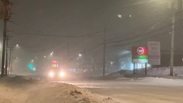 A snowy street early morning during a snowstorm with illuminated streetlights and a car approaching