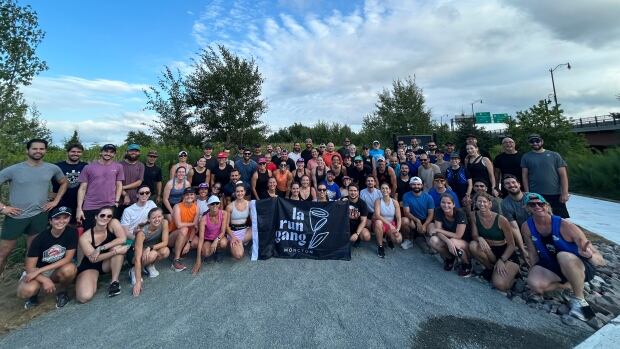 The summers are more popular for La Run Gang and often draw out 60 people each week. The group's busiest summer night brought 80 people to run. A group of around 80 people in summer clothing posing for a group photo. The people in the centre of the photo are holding a black flag that says "la run gang"