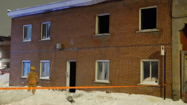 A firefighter stands outside a burnt building surrounded by orange tape. 