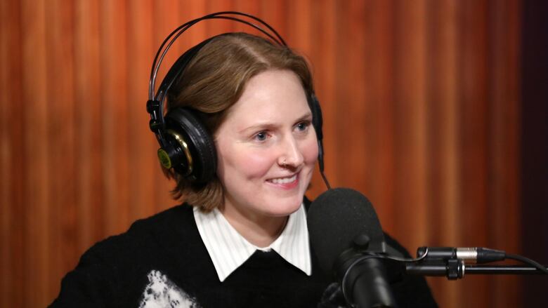 A smiling woman wearing over-ear headphones sits in front of a studio microphone.