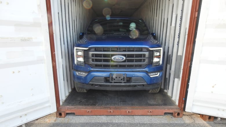 A blue pickup truck inside an open shipping container sitting in a paved space. 