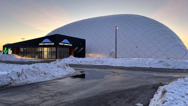 The dome at Credit Union Place, seen near sunset on Wednesday, will be closed indefinitely pending a damage assessment. A large white circular structure is seen against an evening sky behind a building that has an illuminated sign saying Credit Union Place.