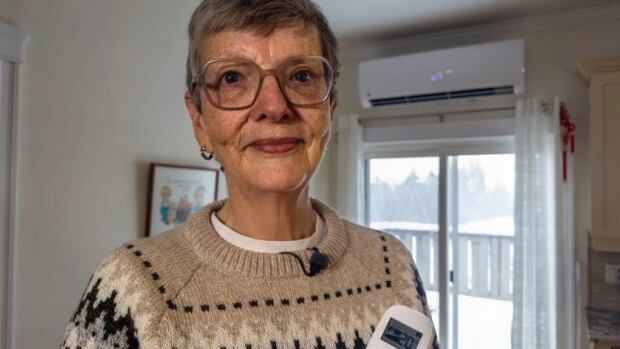 Julie Sentner loves the heat from her two new heat pumps but the cold winter, combined with setting the temperature a bit higher than she used to, means her consumption isn't down as much as she hoped. older woman with short grey hair holding remote for her heat pump which is on the wall of her home in the background