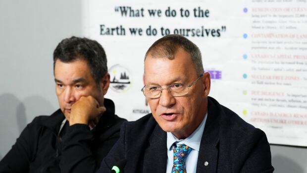 Chief Lance Haymond of Kebaowek First Nation, right, speaks as Algonquin First Nations hold a press conference in Ottawa on Thursday, Aug. 10, 2023., after they were denied a requested for an in-person hearing to make their final presentations to the Canadian Nuclear Safety Commission regarding the proposed new disposal facility at Chalk River. Two mean speak at a press conference. Behind them, a presentation reads, "What we do to the earth we do to ourselves."
