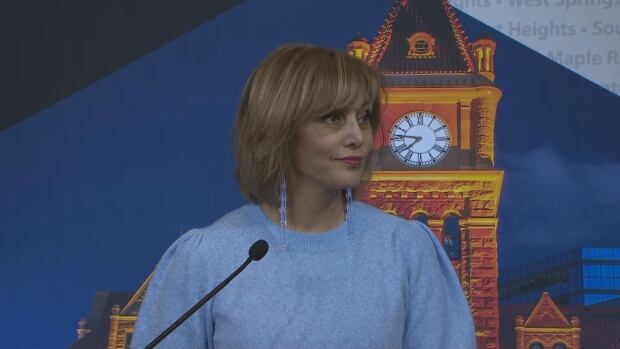 Calgary Mayor Jyoti Gondek has formed a tariff advisory council to shore up economic protections as threats of U.S. tariffs loom. A woman wearing a blue sweater stands in from of a microphone.