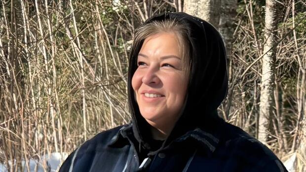 Ramona Nicholas is an Elder and a sweat lodge keeper for Neqotkuk First Nation. A woman stands in nature during the winter, her smiling face turned towards the sun. She wears a blue winter jacket with a plaid pattern, the hood up.