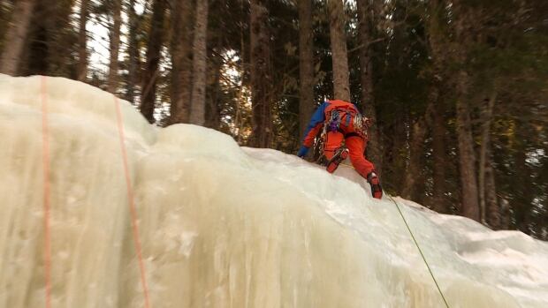 Ice climbing is different from rock climbing, Heidi MacLoud said, because you don't have to search for a route; 'Ice climbing is more of a repetitive motion. You’re always swinging the same,' she said. A person dressed in winter gear and equipped with climbing tools reaches the top of a frozen waterfall in the woods.