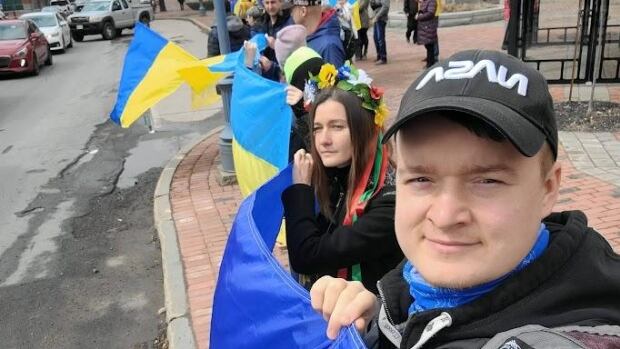 Volodymyr Komarov attends a Ukrainian rally in Moncton. He says Trump's comments about his home country have him feeling angry on the third anniversary of the war. Man holds a Ukrainian flag and stands in a line with other people doing the same.