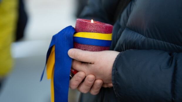 An attendee holds a candle wrapped in the blue and yellow colours of Ukraine at a vigil for victims of the war in Ukraine at the Peace Bridge, in Calgary, AB on March 24, 2022. hands hold a candle. there is a blue and yellow ribbon wrapped around the candle.