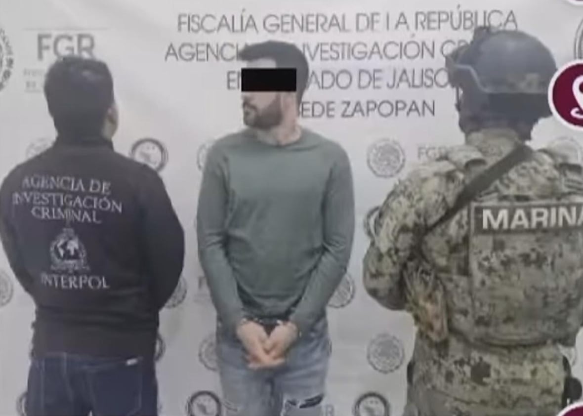 A man in handcuffs is surrounded by someone in an INTERPOL jacket and a soldier in camouflage