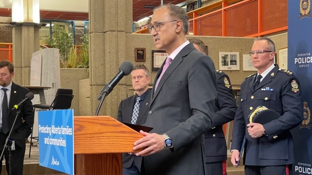 Edmonton mayor Amarjeet Sohi, pictured, said this is a crisis decades in the making and that it will take more than one initiative to solve. Mayor Amarjeet Sohi stands at a podium with a blue sign on the front of it. The sign reads: "Protecting Alberta families and communities." Sohi is wearing a dark grey or black suit with a light pink tie, as well as a smartwatch on his left hand. We are seeing him in profile.
