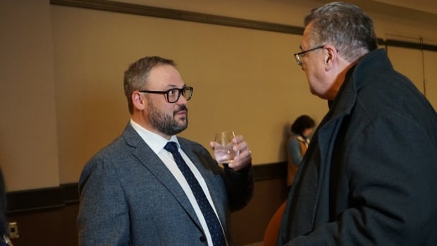 A man with glasses holding a glass while he speaks to another man.