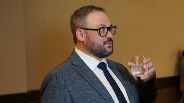 A man with glasses holding a glass while he speaks to another man.