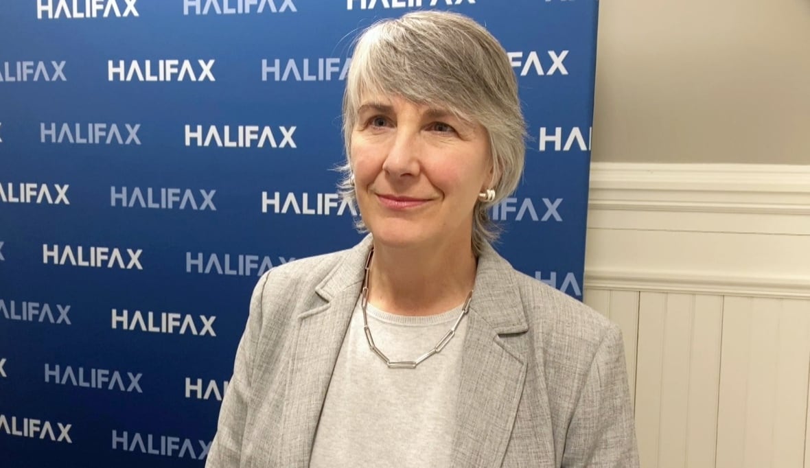 A white woman with short grey hair with bangs in a grey blazer and white shirt stands in front of a blue backdrop featuring the word HALIFAX.