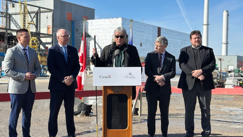 Five people stand at a podium outside near a sewage treatment plant and as two flags blow in the wind behind them.