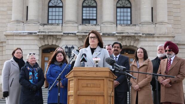 Saskatchewan NDP Leader Carla Beck holds a press conference in front of the legislative building in Regina, Sask., on March 4, 2025. The Saskatchewan NDP is calling for an emergency legislative session in response to tariffs levied by the American government. A woman wearing a grey coat stands at a lectern. She is surrounded by other people wearing outdoor coats.