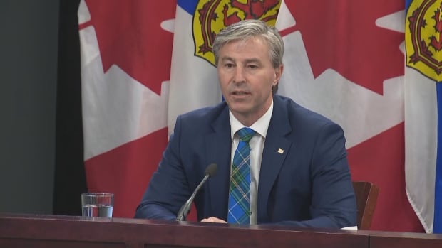 Tim Houston talks to reporters about interprovincial trade barriers on March 5, 2025 in Halifax. A man in a tartan tie sits with Canadian and Nova Scotian flags behind him.