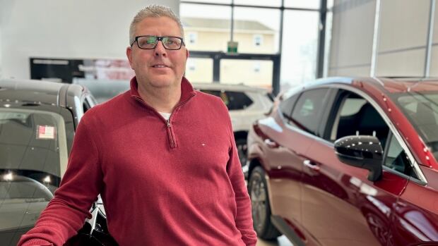Man wears red quarter zip sweater and rests his arm on a car inside of a dealership showroom.