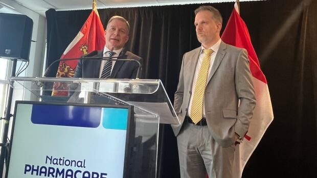 Two mean wearing suits stand behind a podium that says 'national pharmacare.'