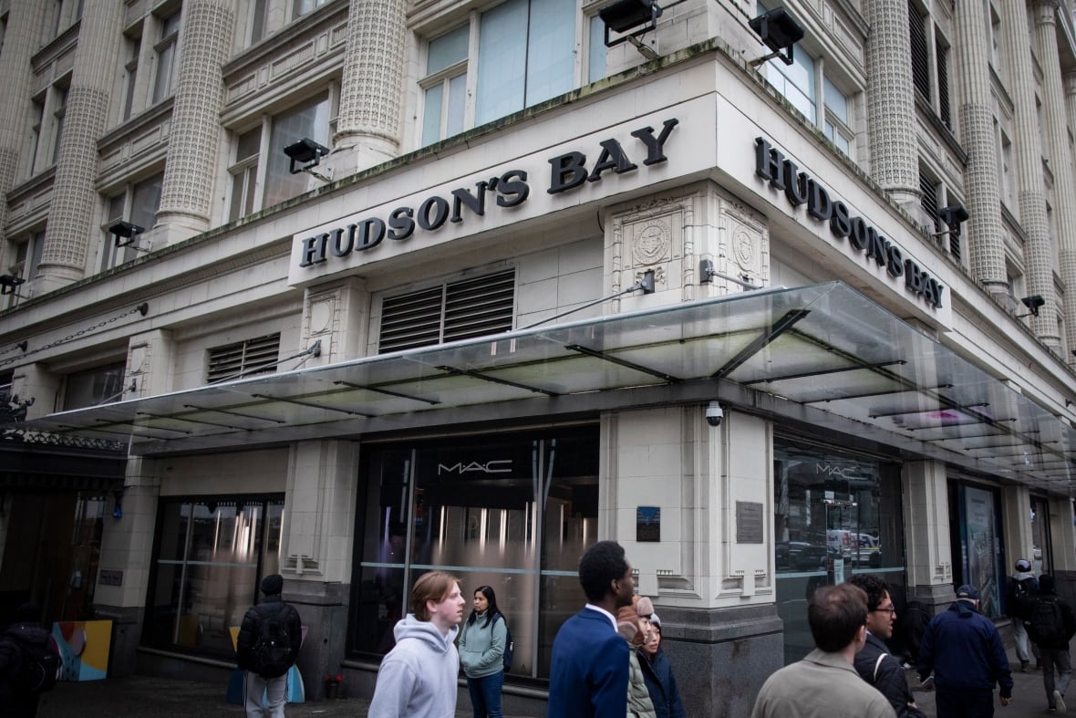 The facade of a large, old columned white building with the words, Hudson's Bay emblazone across it in black letters on the corner of a busy urban street.
