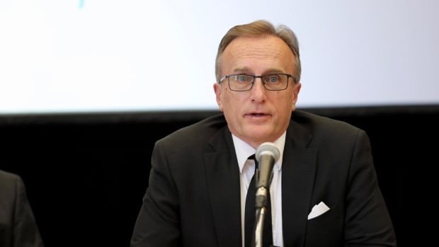 An older man in a black suit and tie sit before a microphone.