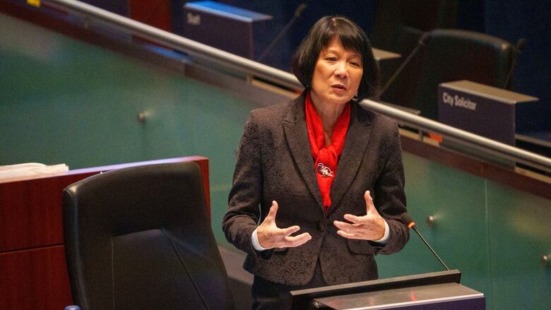 Mayor Olivia Chow stands at her desk in Toronto's city council chambers and speaks into a microphone
