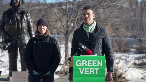 Federal Green Party leader Jonathan Pedneault, right, campaigns in Whitehorse with the party's local candidate, Gabrielle Dupont, March 31, 2025. A man stands behind a podium with a sign that says "Green" on it, beside a river in winter.