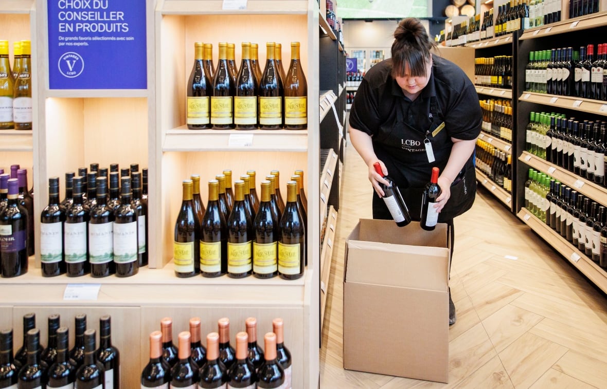 A woman puts wine bottles into a carboard box while standing in a store with bottles on every shelf.