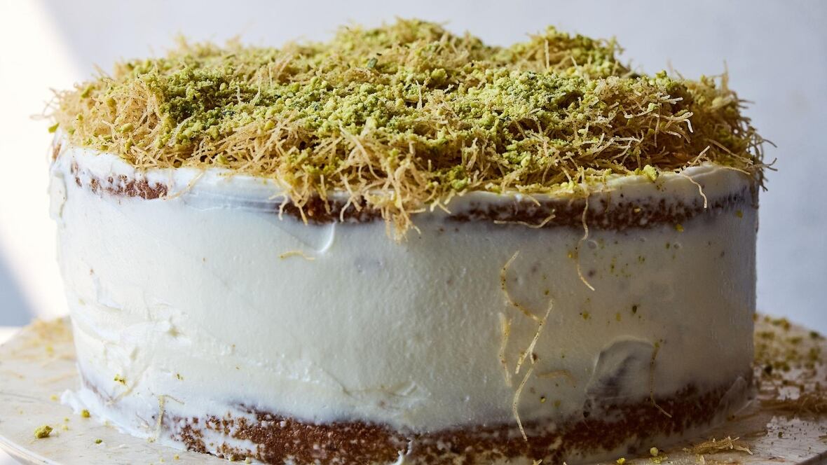 Closeup on a cake with white icing, topped with pistachios and crispy strands of kataifi. It's sitting on a beige marble plate, with a grey background. 