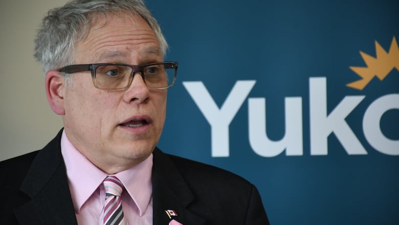 A man with glasses, a pink dress shirt and black suit jacket. He's in front of a sign that says "Yukon." 