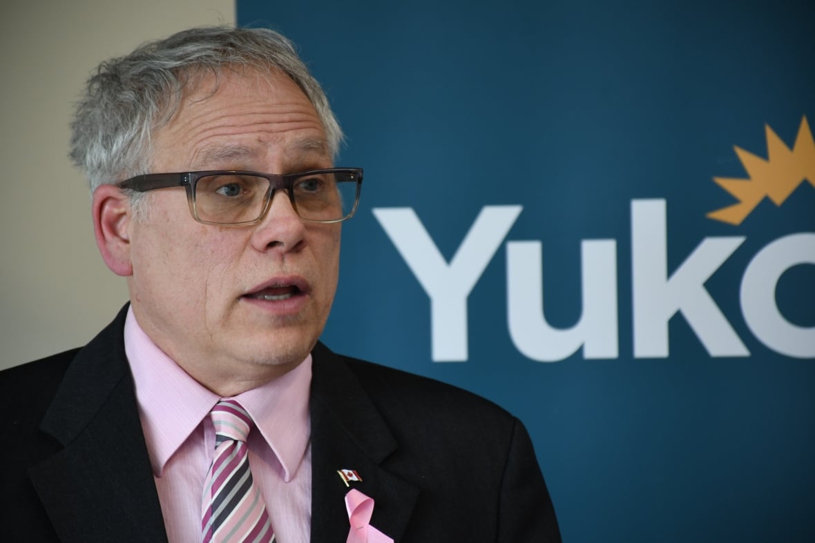 A man with glasses, a pink dress shirt and black suit jacket. He's in front of a sign that says "Yukon." 