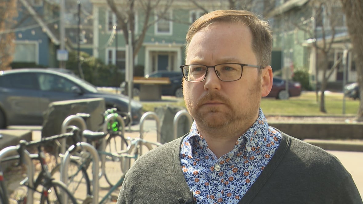 A man with a beard and glasses wears a dark sweater and a floral button-up shirt.