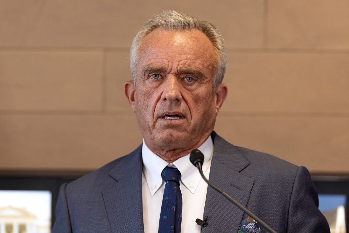 An old man is shown from the shoulders up, looking at the camera with his mouth slightly open. A microphone is in front of him. 