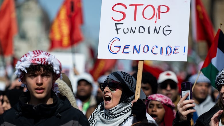 A woman wearing a keffiyeh is seen in a crowd of people holding a sign which reads "Stop Funding Genocide."