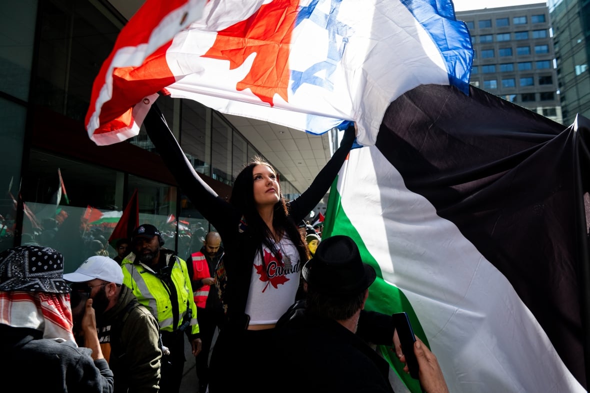A white woman on a crowded street holds up a flag that is half the Canadian flag and half the Israel flag.